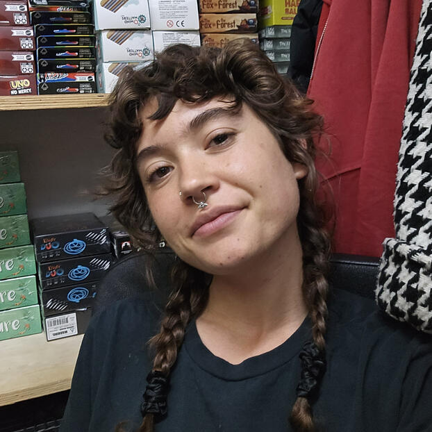 Corvus Corvus, a person with light skin, braided curly brown hair, and a calm smile. The image is of their head, slightly tilted, looking at the camera.
